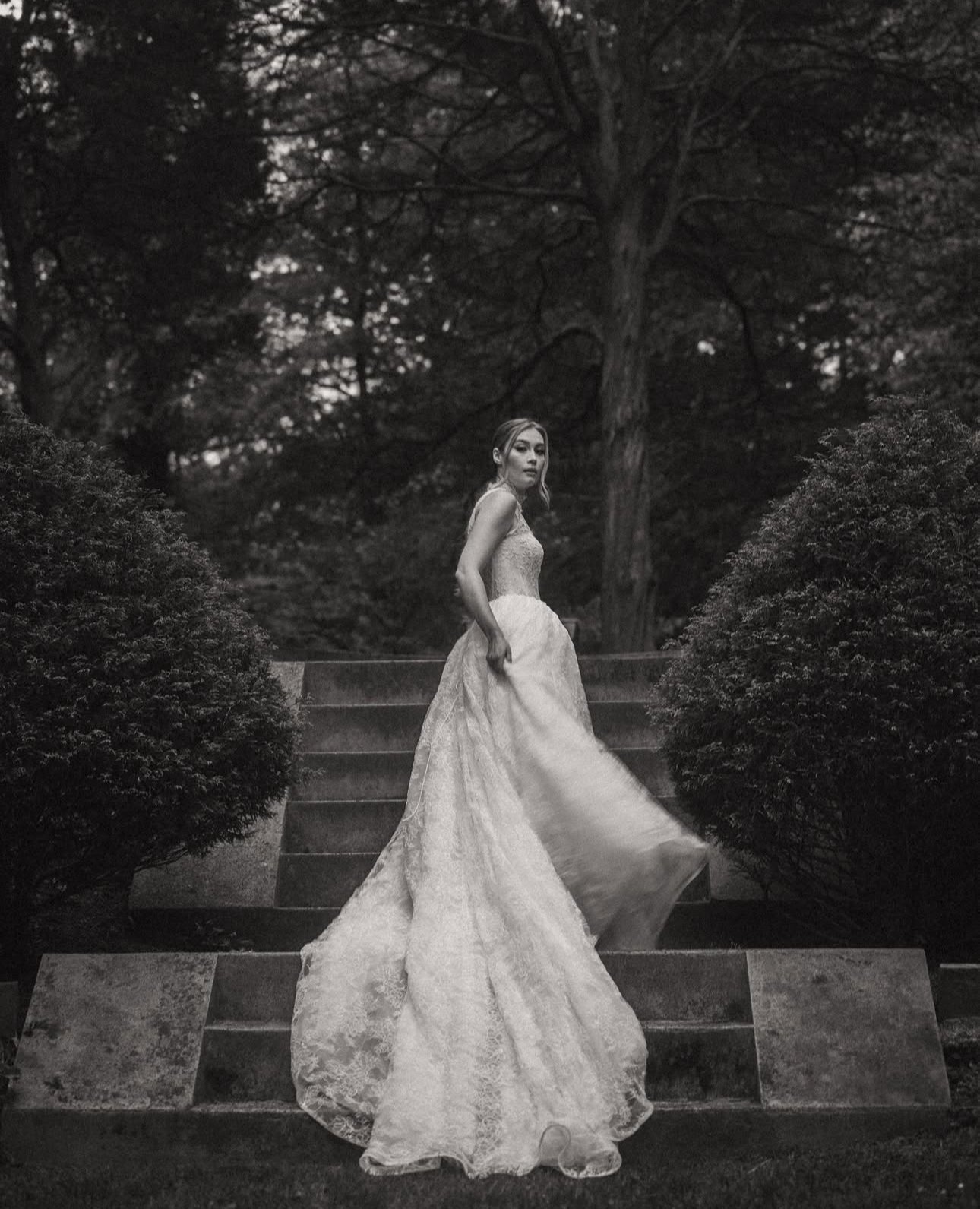 A woman in a wedding dress standing on outdoor steps surrounded by bushes and trees, black and white photo.